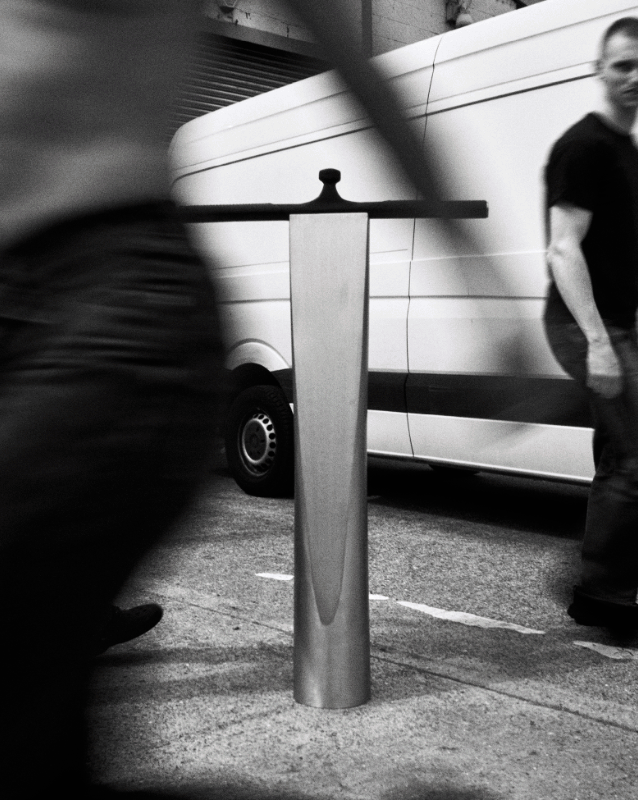 A polished metal sculpture standing on a London pavement, with a van and passers-by blurred behind it.